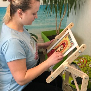 Op de afbeelding: Een vrouw zit op een stoel en gebruikt een houten borduurstandaard om aan een kruissteekproject te werken. De standaard heeft een kantelbaar bovenblad en een verstelbare hoogte. Het borduurproject is een kleurrijk beeld van een religieus icoon.