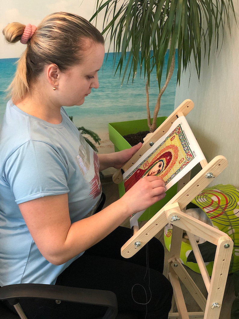 Op de afbeelding: Een vrouw zit op een stoel en gebruikt een houten borduurstandaard om aan een kruissteekproject te werken. De standaard heeft een kantelbaar bovenblad en een verstelbare hoogte. Het borduurproject is een kleurrijk beeld van een religieus icoon.