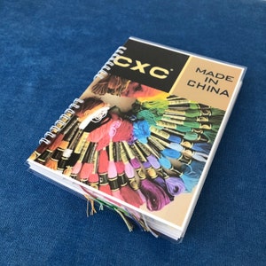 May include: A spiral-bound CXC embroidery floss color chart. The cover features the CXC logo and the text "MADE IN CHINA". The chart displays a wide array of colorful embroidery floss, with strands of floss extending from the pages.