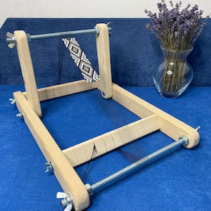 May include: A wooden loom with a black and white beaded pattern being woven on it. The loom has a metal rod and adjustable tension knobs.