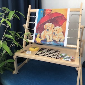 Può includere: Un telaio da ricamo in legno con una tela tesa sopra. La tela presenta un'immagine colorata di due cuccioli sotto un ombrello. Il telaio ha un vassoio sottostante per riporre gli accessori, tra cui un piccolo contenitore di perline e un piccolo contenitore di plastica.