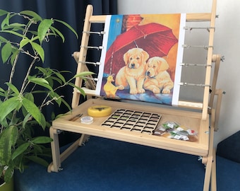 May include: A wooden embroidery stand with a canvas stretched across it. The canvas features a colorful image of two puppies under an umbrella. The stand has a tray underneath for storing supplies, including a small container of beads and a small container of plastic.