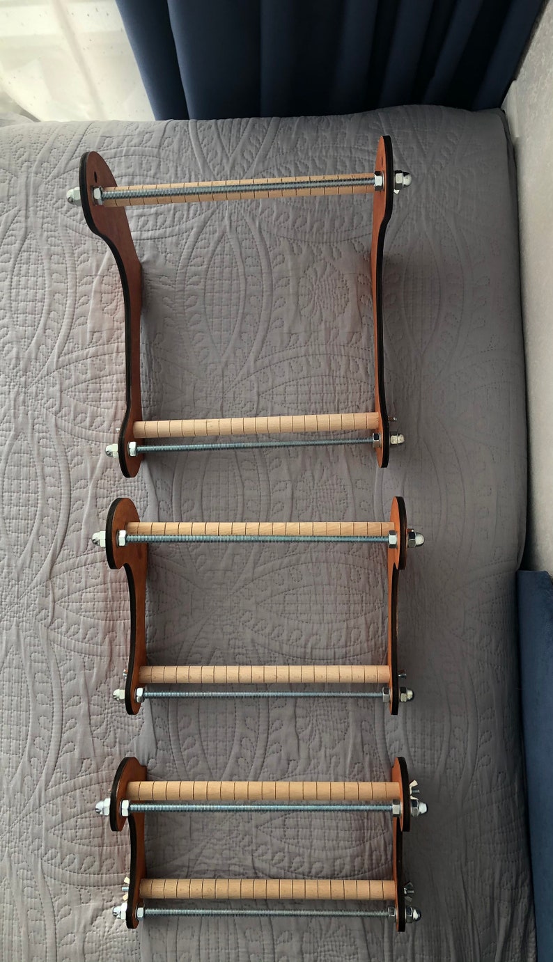 May include: Three wooden loom frames with metal rods and knobs. The frames are stacked on top of each other and are made of light brown wood.