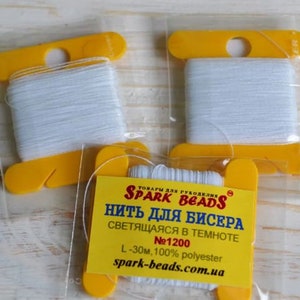 May include: Three spools of white glow-in-the-dark thread for beading, packaged in clear plastic with yellow plastic spools. The packaging has the text "Spark Beads" and "Thread for Beads" in black and red lettering.