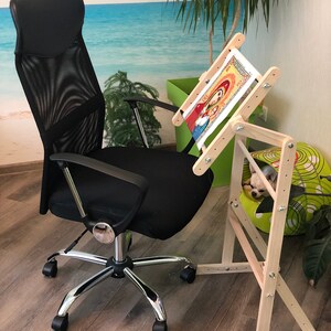 Op de afbeelding: Een houten borduurstandaard met een lopend kruissteekproject. De standaard is verstelbaar en heeft een ingebouwd frame om de stof strak te houden. De standaard staat op een houten vloer met een zwarte bureaustoel op de achtergrond.