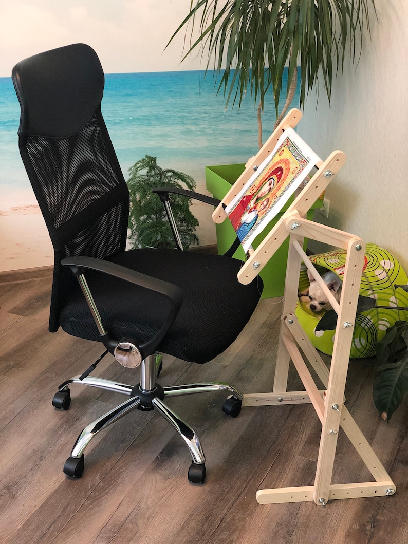 Op de afbeelding: Een houten borduurstandaard met een lopend kruissteekproject. De standaard is verstelbaar en heeft een ingebouwd frame om de stof strak te houden. De standaard staat op een houten vloer met een zwarte bureaustoel op de achtergrond.