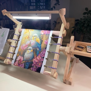 Puede incluir: Un bastidor de bordado de madera con un diseño de pájaros coloridos. El soporte tiene una luz incorporada y una bandeja para suministros. La tela bordada presenta pájaros con tonos azules, amarillos y rosas, sobre un fondo floral.