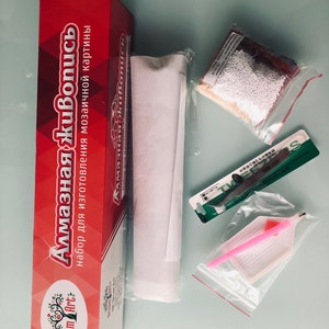 May include: A red box with the text "Алмазная живопись" and "Dream Art" on it. The box contains a white canvas, a bag of white beads, a pink tool, a green tool, and a clear plastic bag with a pink tool.