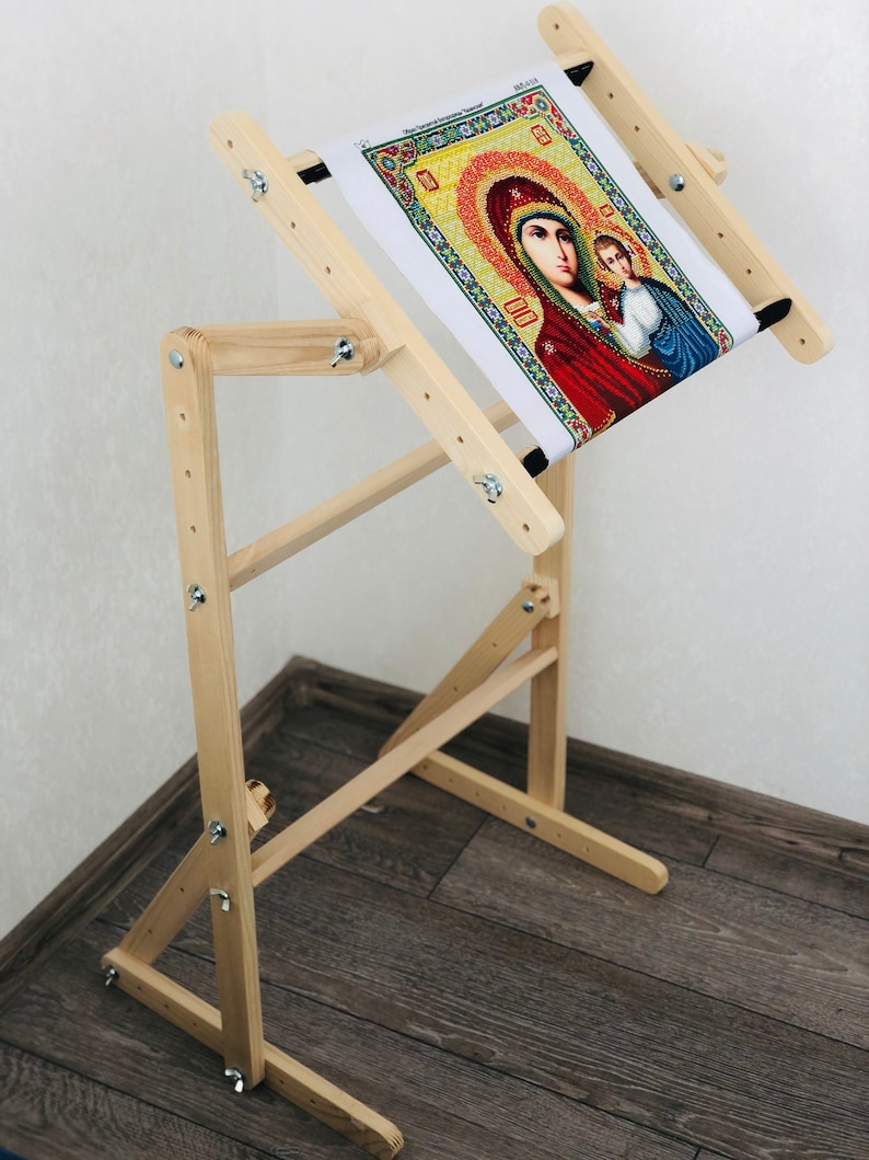 Op de afbeelding: Een houten borduurstandaard met een kantelbaar bovenblad en verstelbare hoogte. De standaard is gemaakt van natuurlijk hout en heeft een donkerbruine afwerking. Een borduurproject met kruissteek is op de standaard te zien.