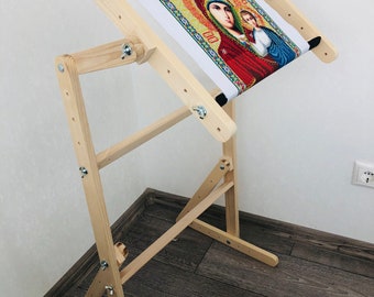 May include: A wooden embroidery stand with a canvas stretched across it. The canvas features a colorful image of a religious icon. The stand has adjustable legs and a built-in frame.