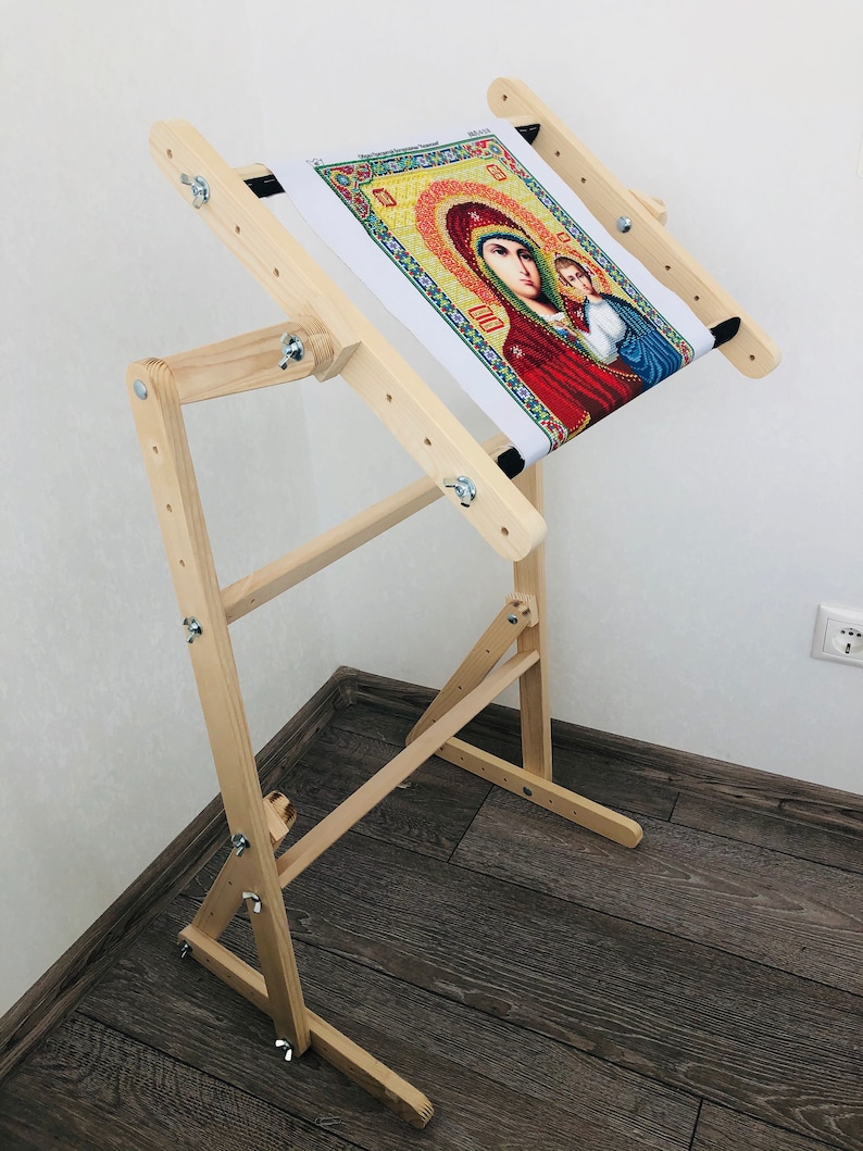 Op de afbeelding: Een houten borduurstandaard met een doek erover gespannen. Het doek heeft een kleurrijk beeld van een religieus icoon. De standaard heeft verstelbare poten en een ingebouwd frame.