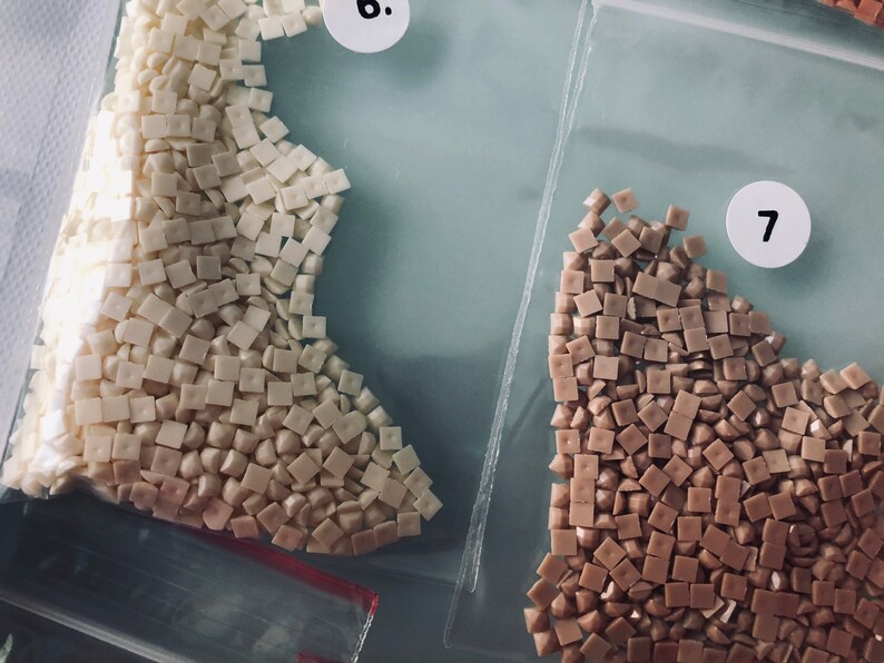 May include: Two small bags of diamond painting drills. The bag on the left contains white drills, and the bag on the right contains brown drills. The bags are labeled with the numbers 6 and 7.