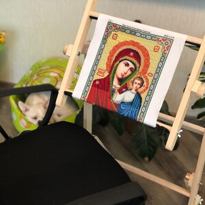 Op de afbeelding: Een houten borduurstandaard met een wit canvas waarop een kleurrijk diamant schilderij van de Maagd Maria en baby Jezus te zien is. De standaard is in hoogte en spanning verstelbaar.