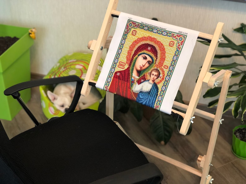 Op de afbeelding: Een houten borduurstandaard met een wit canvas waarop een kleurrijk diamant schilderij van de Maagd Maria en baby Jezus te zien is. De standaard is in hoogte en spanning verstelbaar.