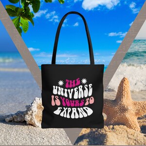 May include: Black tote bag with white text that reads "The Universe Is Yours To Expand" in a retro font. The bag is on a beach with a starfish and seashells.