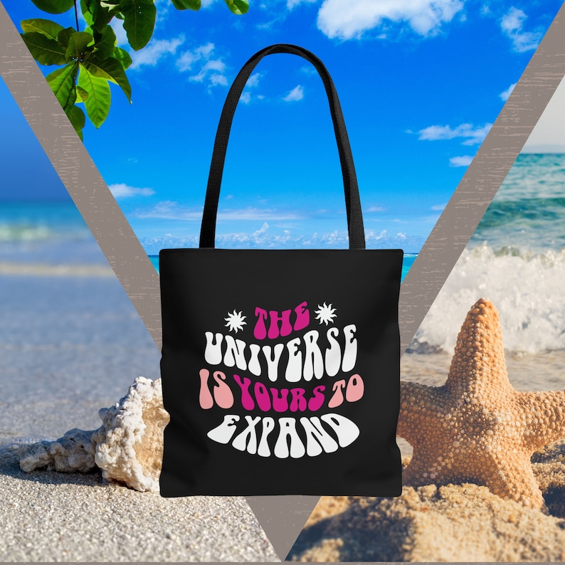 May include: Black tote bag with white text that reads "The Universe Is Yours To Expand" in a retro font. The bag is on a beach with a starfish and seashells.