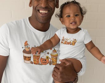 May include: A white t-shirt featuring a graphic of autumnal drinks, including a pumpkin spice latte, a hot chocolate, and a caramel apple cider. The shirt is being worn by a person holding a child wearing a matching white onesie with a smaller version of the same graphic.