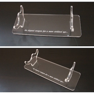 May include: Clear acrylic display stand with a rectangular base and two curved supports. The stand features the engraved text "An elegant weapon for a more civilized age..." Ideal for showcasing collectibles or decorative items.