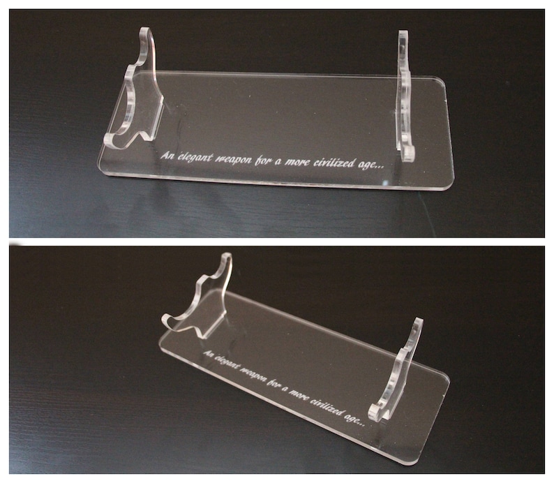 May include: Clear acrylic display stand with a rectangular base and two curved supports. The stand features the engraved text "An elegant weapon for a more civilized age..." Ideal for showcasing collectibles or decorative items.