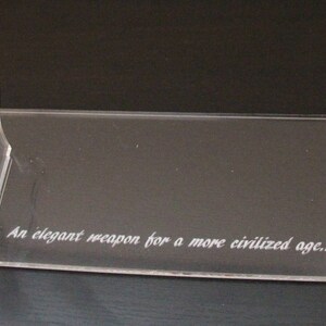 May include: Clear acrylic display stand with a rectangular base and two curved supports. The stand features the engraved text: "An elegant weapon for a more civilized age...". Ideal for displaying collectibles.