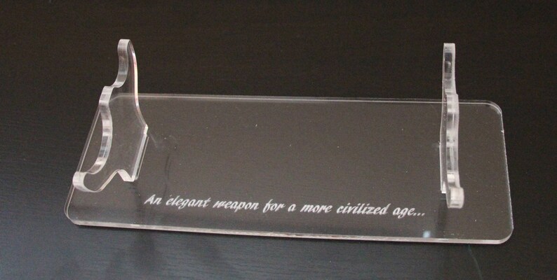 May include: Clear acrylic display stand with a rectangular base and two curved supports. The stand features the engraved text: "An elegant weapon for a more civilized age...". Ideal for displaying collectibles.