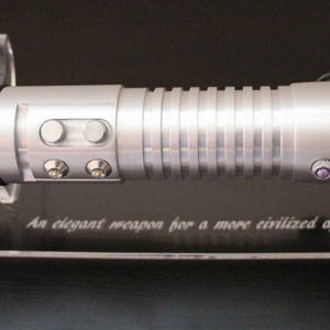 May include: A silver lightsaber with a ribbed handle and multiple buttons, resting on a clear stand. The stand has the text "An elegant weapon for a more civilized age..." etched on it. The lightsaber has black accents and a cylindrical design.