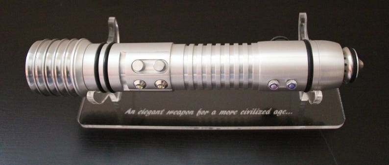 May include: A silver lightsaber with a ribbed handle and multiple buttons, resting on a clear stand. The stand has the text "An elegant weapon for a more civilized age..." etched on it. The lightsaber has black accents and a cylindrical design.