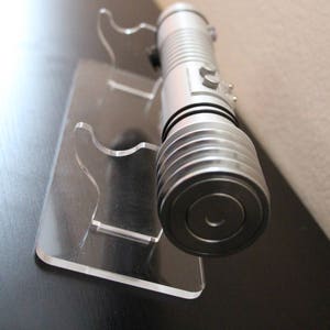 May include: A silver lightsaber rests on a clear acrylic display stand. The lightsaber has a cylindrical design with ribbed sections and a circular end. The stand has a rectangular base and two curved supports to hold the lightsaber.