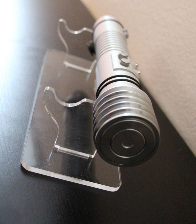 May include: A silver lightsaber rests on a clear acrylic display stand. The lightsaber has a cylindrical design with ribbed sections and a circular end. The stand has a rectangular base and two curved supports to hold the lightsaber.