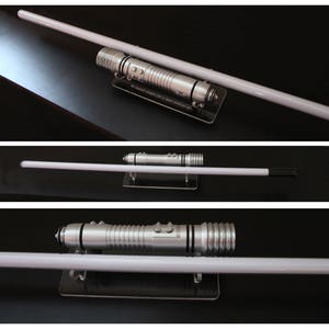 May include: A silver lightsaber with a white blade, displayed on a black surface. The hilt features detailed metallic accents and buttons. The blade is long and cylindrical, with a bright, glowing appearance. The lightsaber is a prop from a science fiction franchise.