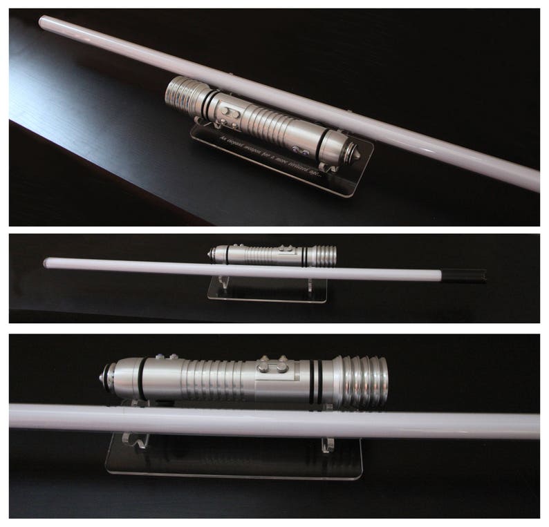 May include: A silver lightsaber with a white blade, displayed on a black surface. The hilt features detailed metallic accents and buttons. The blade is long and cylindrical, with a bright, glowing appearance. The lightsaber is a prop from a science fiction franchise.