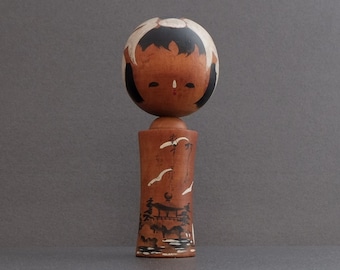 May include: A wooden Kokeshi doll with a round head featuring black hair and a light brown face. The cylindrical body is stained brown and decorated with a black and white painted landscape scene depicting a bridge, trees, and flying birds.