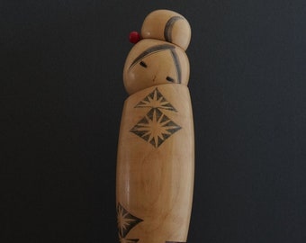 May include: A wooden figurine with a red dot on its head, stacked with three other wooden pieces. Each piece has a geometric star design. The figurine is standing on a red base.