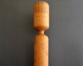May include: A wooden doll with a simple face and a patterned body. The doll is made of light brown wood and has a cylindrical shape.