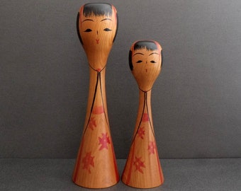 Könnte beinhalten: Zwei Holzpuppen mit bemalten Gesichtern und roten und schwarzen Details. Die Puppen tragen rote und schwarze Kleider mit einem Blatt-Muster. Die größere Puppe ist größer als die kleinere Puppe.