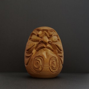 May include: A wooden carving of a Japanese lucky charm, known as a Daruma doll. The doll has a round body with a painted face and a red and white design. The doll is a symbol of good luck and perseverance.