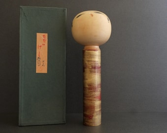 Könnte beinhalten: Eine japanische Holzpuppe mit bemaltem Gesicht und einem rot-goldenen bemalten Körper. Die Puppe steht auf einem Holzsockel und befindet sich in einer grünen Schachtel mit japanischen Schriftzeichen.