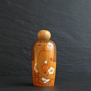 Pode incluir: Boneca Kokeshi japonesa de madeira com cabeça arredondada e corpo cilíndrico. A boneca é pintada com uma cor de base castanho claro e apresenta um desenho floral com flores brancas e vermelhas. O cabelo da boneca é pintado de castanho.