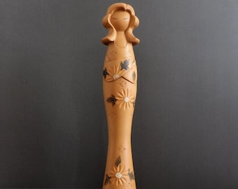 May include: A wooden figurine of a woman with long brown hair, wearing a carved floral dress. The figurine is painted in shades of brown and black, with white accents.
