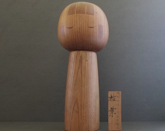 May include: A wooden figurine of a person with closed eyes. The figurine is made of light brown wood and has a simple, minimalist design. The figurine is standing on a gray surface. The text "松寿" is written on a small wooden plaque next to the figurine.