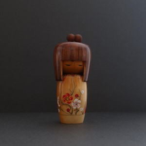 Könnte beinhalten: Eine japanische Holzpuppe Kokeshi mit braunen Haaren, geschlossenen Augen und einem bemalten Blumenmuster auf ihrem Kleid.