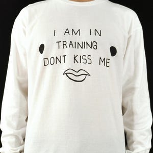 May include: A white long-sleeve shirt featuring black hand-drawn style text that reads I AM IN TRAINING DONT KISS ME, accompanied by two black dots on either side and a simple line drawing of lips centered below the text on the chest area.