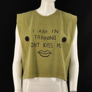 May include: Olive green sleeveless crop top with the text "I AM IN TRAINING ONT KISS ME" in black. The shirt features two black circles and a black lip graphic. The top is displayed on a mannequin.