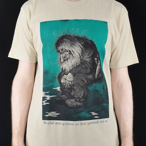 May include: A beige t-shirt with a green background image of a troll sitting on a rock. The text "Trollst som grunner på hvor gammelt det er" is printed below the image.