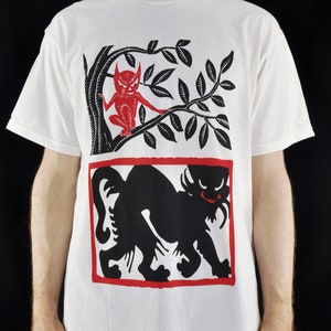 May include: White t-shirt with a black and red graphic print of a black cat and a red devil in a tree.