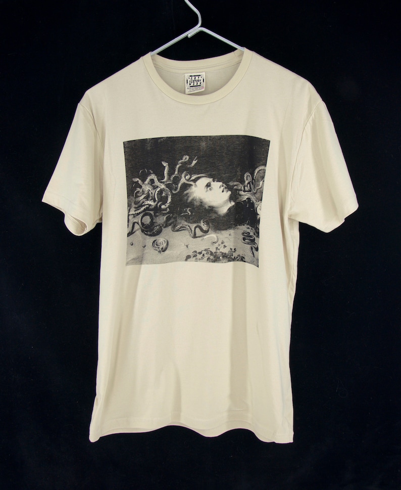 K&ouml;nnte beinhalten: Ein cremefarbenes T-Shirt mit einem schwarz-wei&szlig;en Bild von Medusa, einer mythischen griechischen Figur mit Schlangenhaaren, das auf der Vorderseite aufgedruckt ist. Das Bild ist von einem schwarzen Rahmen umgeben.