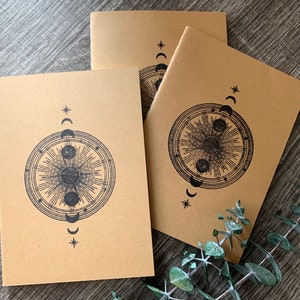 May include: Three brown kraft paper notebooks with a black and white moon and star design. The design features a circle with a sun and moon inside, surrounded by a ring of stars and moons.