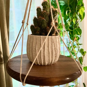 May include: A hanging plant shelf made of dark wood with a white ceramic pot containing a green cactus plant. The shelf is suspended by a macrame rope.