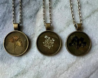 May include: Three antique bronze-toned pendant necklaces. Each pendant features a circular glass face with a pressed flower design. The flowers are in shades of brown, white, and black. The necklaces hang on delicate bronze chains.