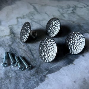 May include: Four silver-colored, hammered-texture cabinet knobs with matching screws. The knobs are round with a slightly domed shape. The hardware is displayed on a marble-patterned surface.